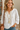 Woman wearing a white embroidered blouse with a necklace, standing indoors.